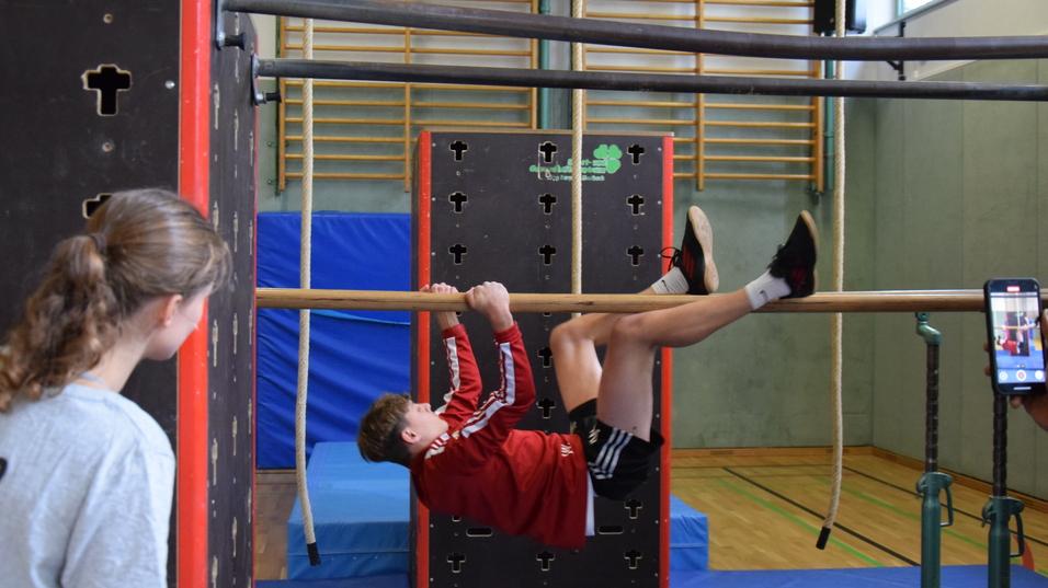 Junge &uuml;bt am Stangenparcours, w&auml;hrend ein M&auml;dchen zuschaut, im Hintergrund eine Turnhalle.