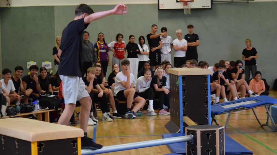 Junge balanciert auf einem Balken, w&auml;hrend Zuschauer in einer Sporthalle zuschauen.