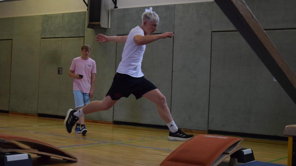 Jugendlicher springt &uuml;ber Hindernisse in einer Sporthalle, w&auml;hrend ein weiterer Zuschauer im Hintergrund steht.
