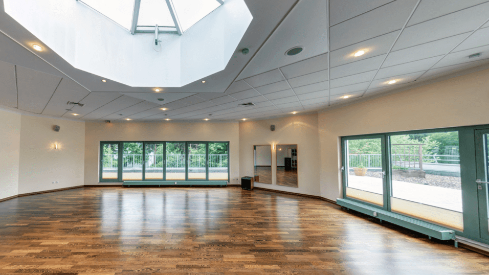 Heller Veranstaltungsraum mit Holzfu&szlig;boden, gro&szlig;en Fenstern und Oberlicht. Ein Spiegel und T&uuml;ren sind sichtbar.