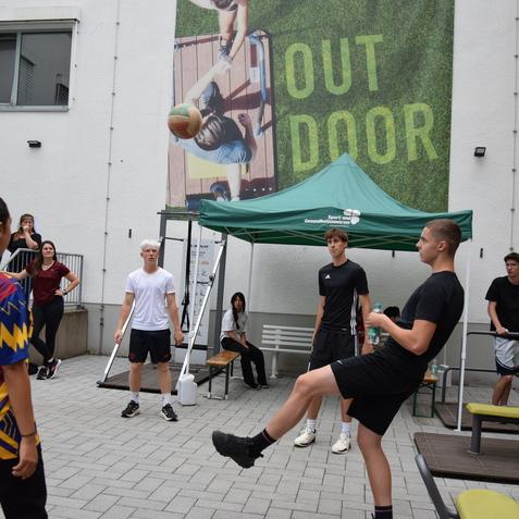 Gruppenspiel in einem Innenhof mit Jugendlichen, einem Zelt und einer Wand mit „OUTDOOR“-Grafik im Hintergrund.