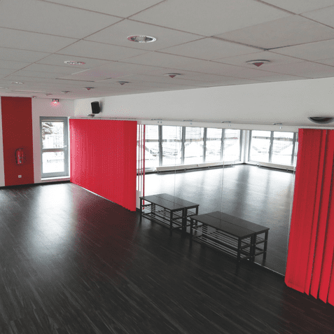 Leerer Raum mit Holzfußboden, großen Fenstern, Spiegelwand und roten Vorhängen. Zwei schwarze Tische stehen in der Mitte.