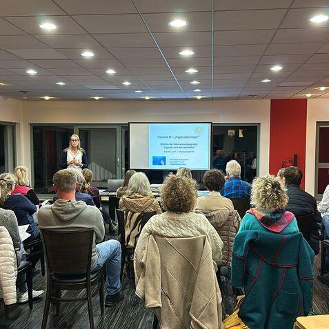 Vortragende pr&auml;sentiert vor Publikum in einem Raum mit Bildschirmen und St&uuml;hlen, Teilnehmer sitzen in Reihen.