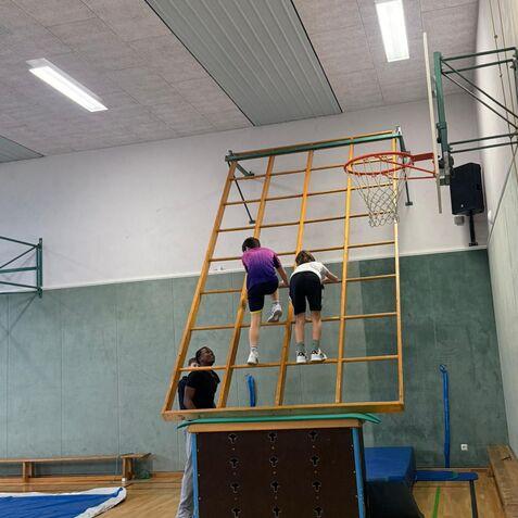 Zwei Kinder klettern an einer Holzleiter, während ein Erwachsener sie im Hintergrund unterstützt.