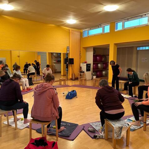 Gruppe von Erwachsenen sitzt auf Stühlen in einem bunten Raum, während sie an einer Übung oder Aktivität teilnehmen.