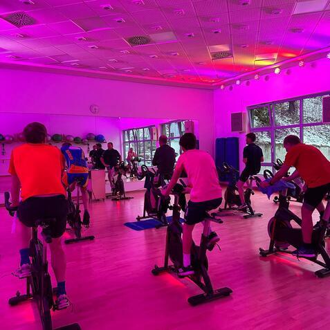 Spinning-Kurs mit mehreren Teilnehmern in einem gut beleuchteten Raum mit pinker ambiente Beleuchtung.