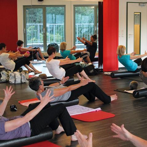 Gruppentraining im Yoga-Studio mit Teilnehmern, die auf Matten &Uuml;bungen ausf&uuml;hren, umgeben von gro&szlig;en Fenstern.