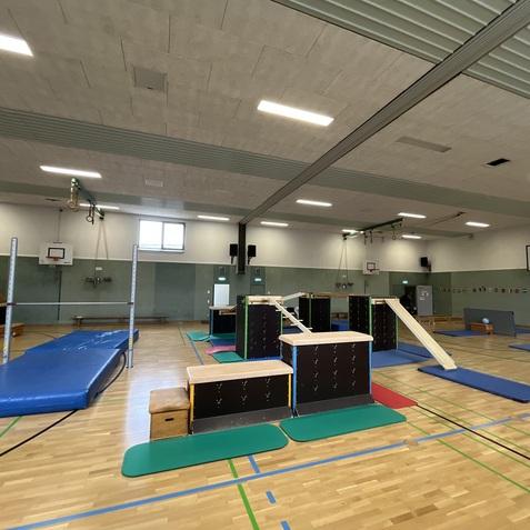 Turnhalle mit verschiedenen Ger&auml;ten, darunter eine Matte, Barren und Kasten, auf einem Holzfu&szlig;boden.