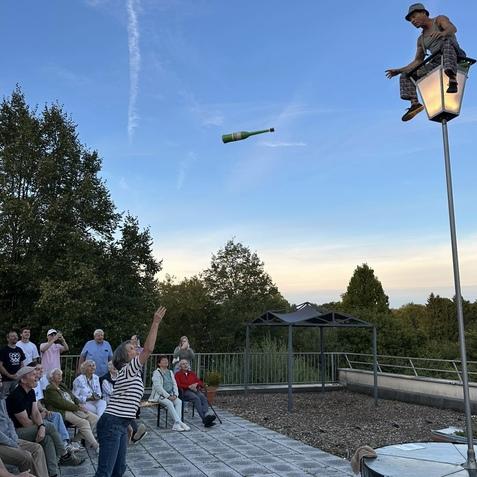 Eine Person wirft eine Flasche zu einem Balancierer auf einer Laterne, w&auml;hrend eine Gruppe Zuschauer zusieht.