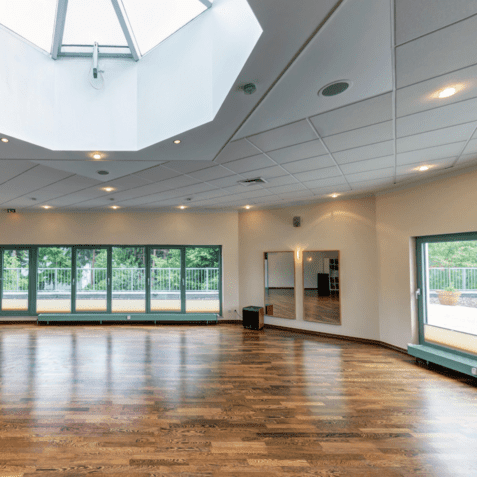 Heller Veranstaltungsraum mit Holzfußboden, großen Fenstern und Oberlicht. Ein Spiegel und Türen sind sichtbar.