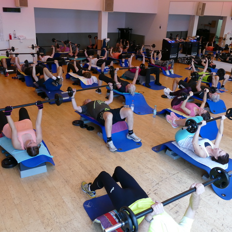 Gruppe von Sportlern beim Bodypump-Training auf blauen B&auml;nken in einem Fitnessstudio, Gewichte werden lifted.