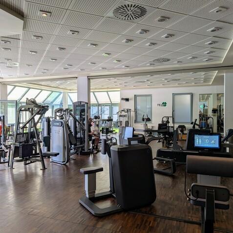 Fitnessstudio mit verschiedenen Trainingsgeräten und großen Fenstern, die Tageslicht hereinlassen.