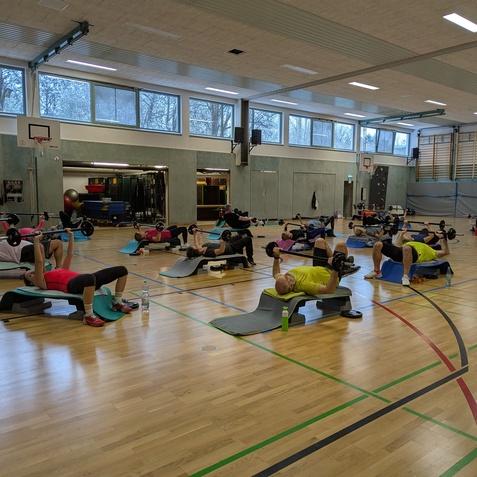 Gruppe von Personen beim Krafttraining auf Matten in einer Sporthalle, umgeben von Fitnessgeräten.