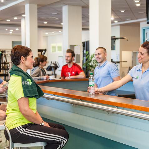 Fitnessstudio mit G&auml;sten an der Theke; eine Angestellte &uuml;berreicht einer Frau in Sportkleidung eine Wasserflasche.
