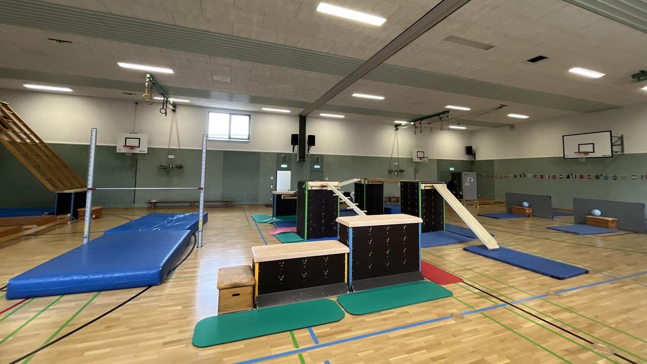 Turnhalle mit verschiedenen Ger&auml;ten, darunter eine Matte, Barren und Kasten, auf einem Holzfu&szlig;boden.
