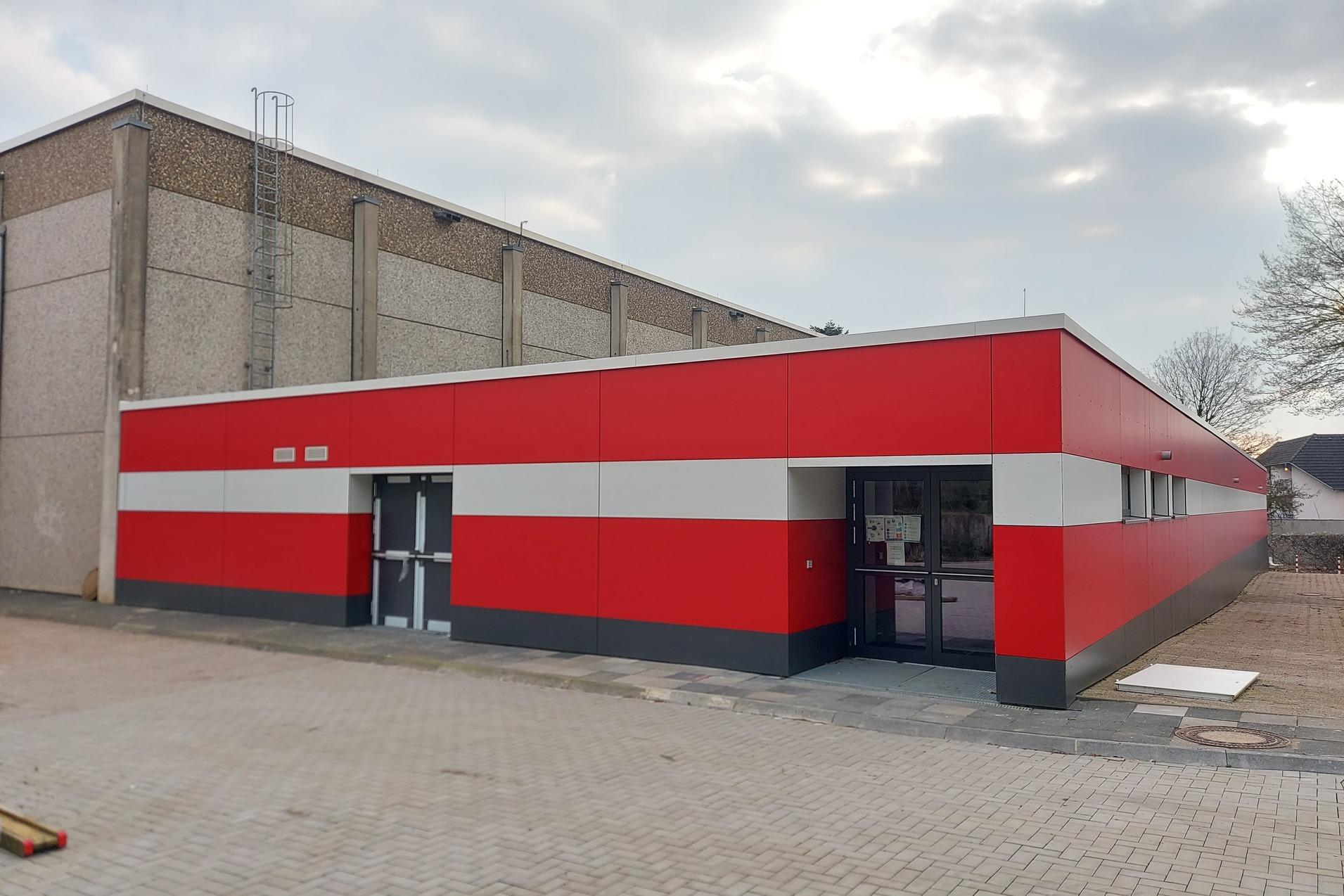 Neubau eines flacheren Gebäudes mit roten und weißen Streifen, angrenzend an ein höheres, graues Gebäude.