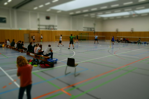 Badminton-Spieler auf einem Court in einer Sporthalle, Zuschauer sitzen auf B&auml;nken entlang des Spielfelds.
