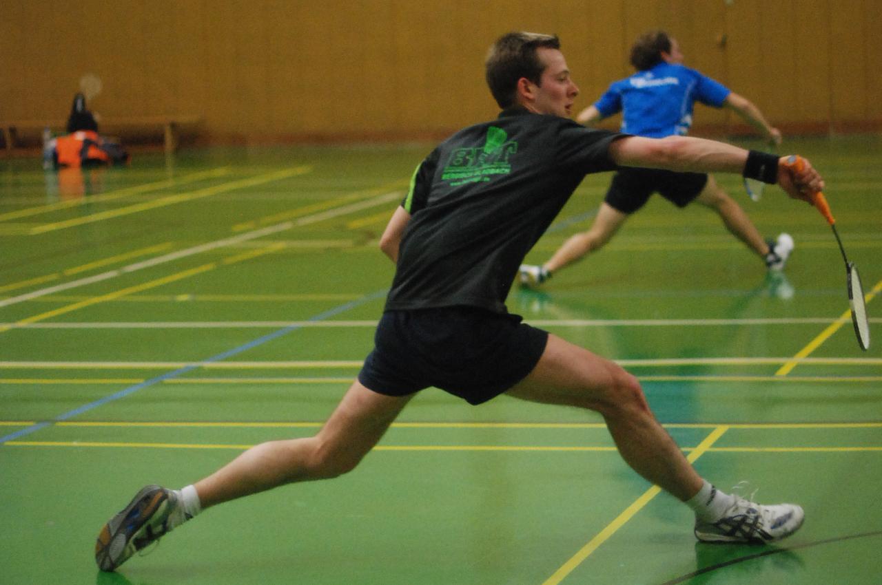 Zwei Badmintonspieler in Aktion auf einem gr&uuml;nen Spielfeld, einer schl&auml;gt mit einem Schl&auml;ger einen Federball.