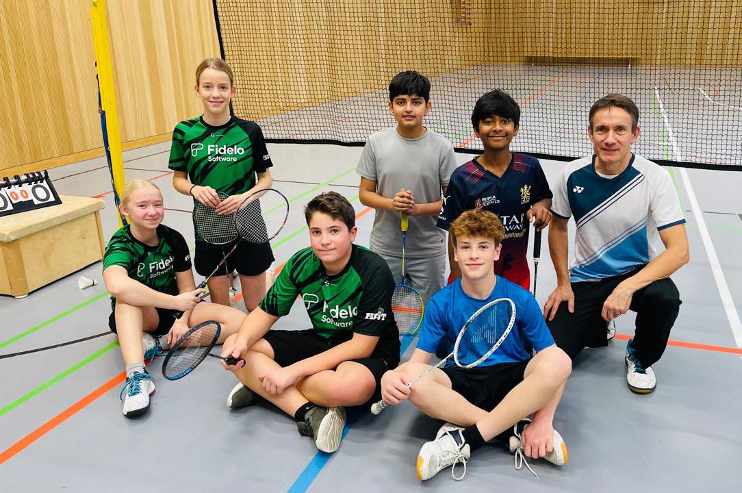 Sechs Kinder und ein Trainer posieren in Sportkleidung mit Badmintonschl&auml;gern in einer Halle. Hintergrund mit Netz und Holzw&auml;nden.