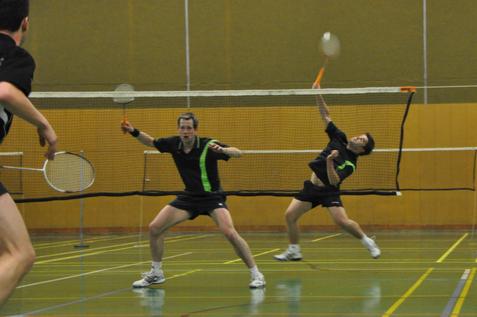Zwei Badmintonspieler in Aktion, einer springt, der andere bereit zum Schlag, Netz im Hintergrund.