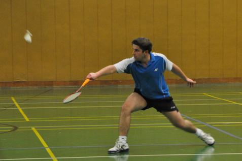 Ein Badmintonspieler in Aktion, der einen Federball mit einem Schl&auml;ger in einer Sporthalle schl&auml;gt.
