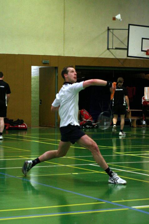 Ein Badmintonspieler schl&auml;gt einen Federball in einer Sporthalle mit gr&uuml;nem Boden.