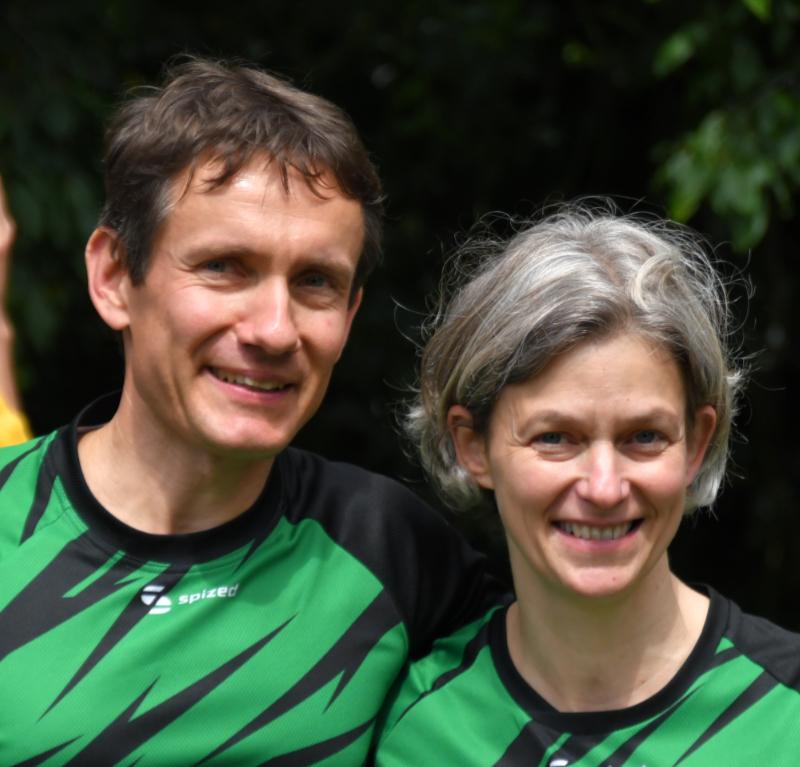 Portrait eines Mannes und einer Frau in gr&uuml;nen Sporttrikots, beide l&auml;cheln vor einer gr&uuml;nen Hintergrundlandschaft.