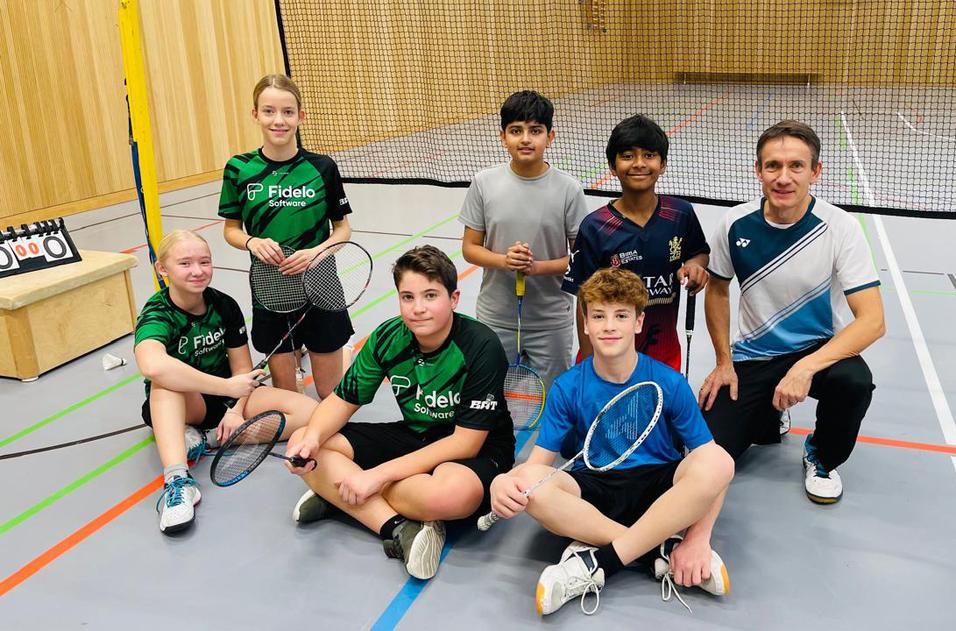 Sechs Kinder und ein Trainer posieren in Sportkleidung mit Badmintonschl&auml;gern in einer Halle. Hintergrund mit Netz und Holzw&auml;nden.