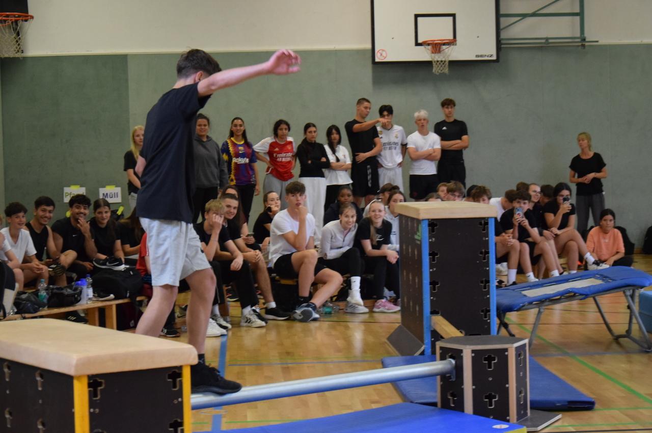 Junge balanciert auf einem schmalen Balken, umgeben von zuschauenden Sch&uuml;lern in einer Sporthalle.