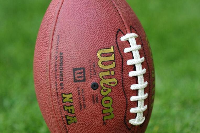 Ein American Football steht auf einem schwarzen Tee, mit gr&uuml;nem Rasen im Hintergrund.