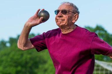 &Auml;lterer Mann mit Sonnenbrille wirft eine Kugel im Freien auf einem Sportplatz. Hintergrund mit Fu&szlig;balltor und B&auml;umen.