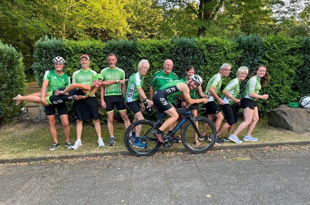 Gruppe von zehn Radfahrern in grünen Trikots posiert fröhlich im Freien, einige halten Fahrräder, andere lachen.