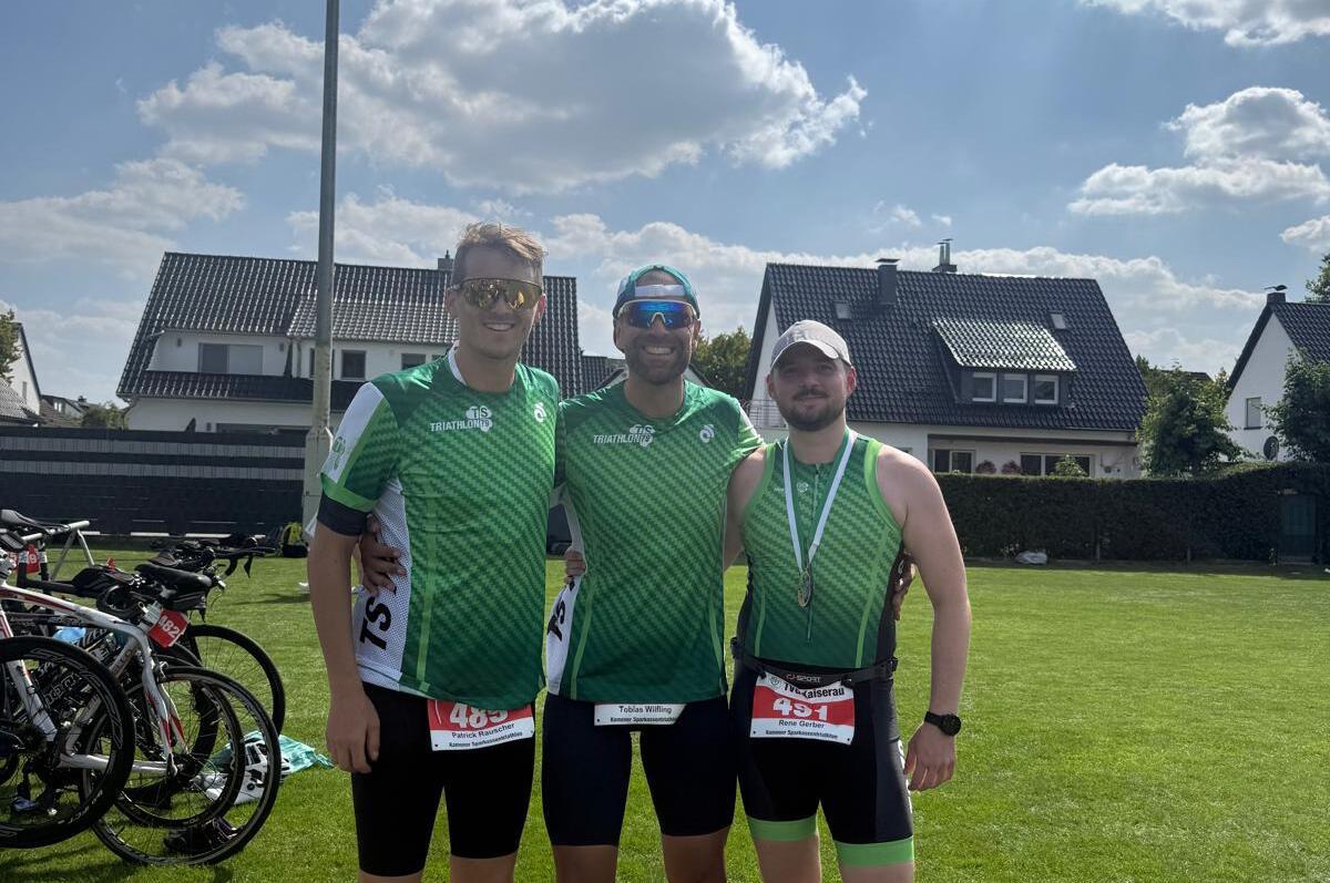 Drei Athleten in grünen Trikots mit Medaille stehen im Gras, im Hintergrund Fahrräder und ein blauer Himmel.