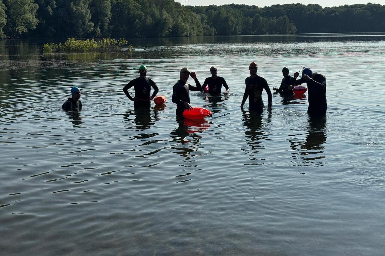 Gruppen von Schwimmern in Neoprenanzügen stehen im Wasser eines ruhigen Sees, einige halten orangefarbene Schwimmbojen.
