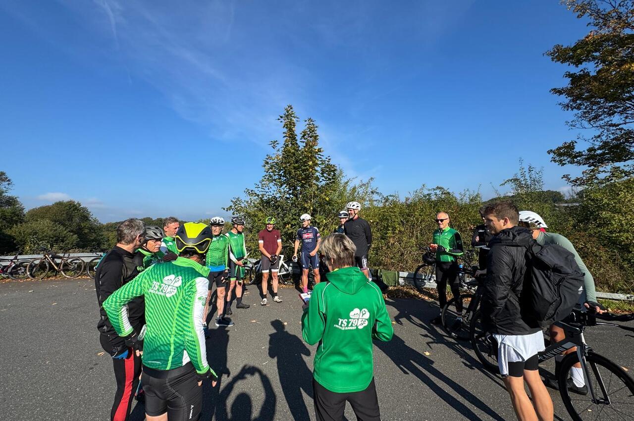 Gruppe von Radfahrern in grünen Trikots versammelt sich zur Besprechung unter blauem Himmel.