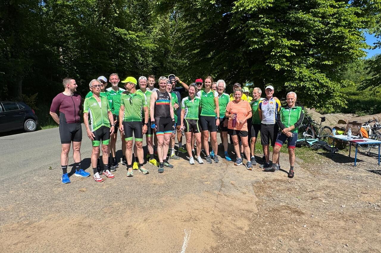 Gruppe von Radfahrern in grünen Trikots posiert im Freien vor Bäumen und einem Fahrradparkplatz.