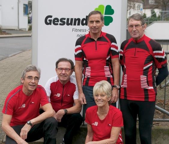 Fünf sportlich gekleidete Personen in roten Shirts posieren vor einem Fitnesscenter-Schild.