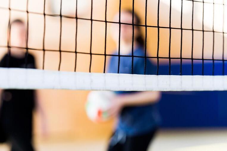 Netz eines Volleyballfeldes mit unscharfen Spielern im Hintergrund, einer h&auml;lt einen Volleybal.