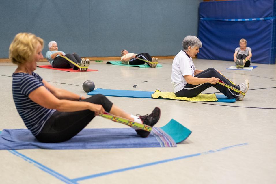 F&uuml;nf Frauen sitzen auf Gymnastikmatten und trainieren mit Widerstandsb&auml;ndern in einem Sportraum.