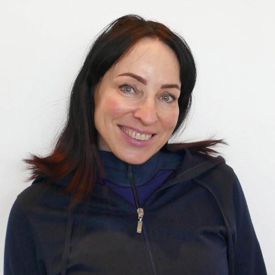 Lächelnde Frau mit langen, dunklen Haaren in einem dunklen Sweatshirt vor weißem Hintergrund.