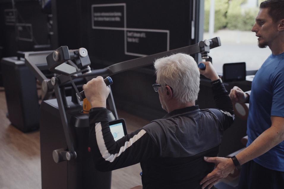 Mann mit grauen Haaren trainiert an einer Fitnessmaschine, während ein Trainer ihn anleitet und unterstützt.