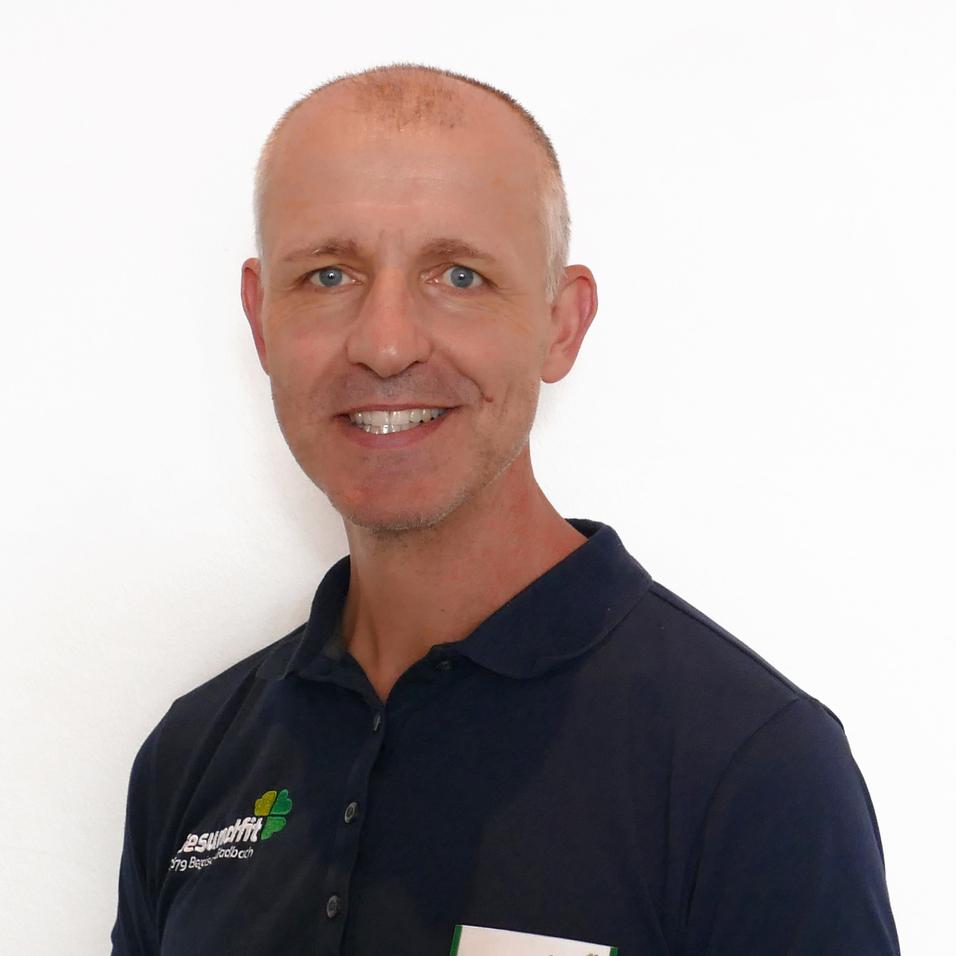 Lächelnder Mann in einem schwarzen Poloshirt mit Namensschild vor einem weißen Hintergrund.