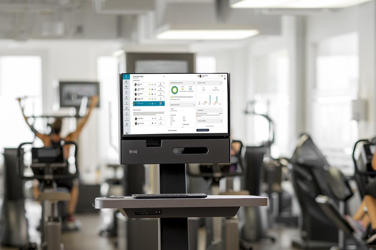 Fitnessstudio mit einem monitorisierten Fitnessger&auml;t auf einem stabilen Tisch. Im Hintergrund trainierende Personen.