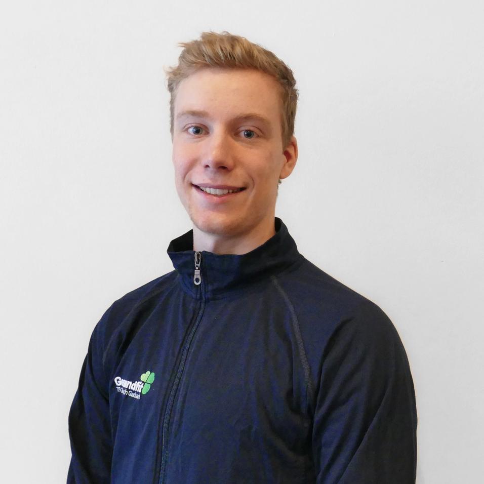 Junger Mann in einem dunklen Trainingsjacke lächelt vor weißer Wand.