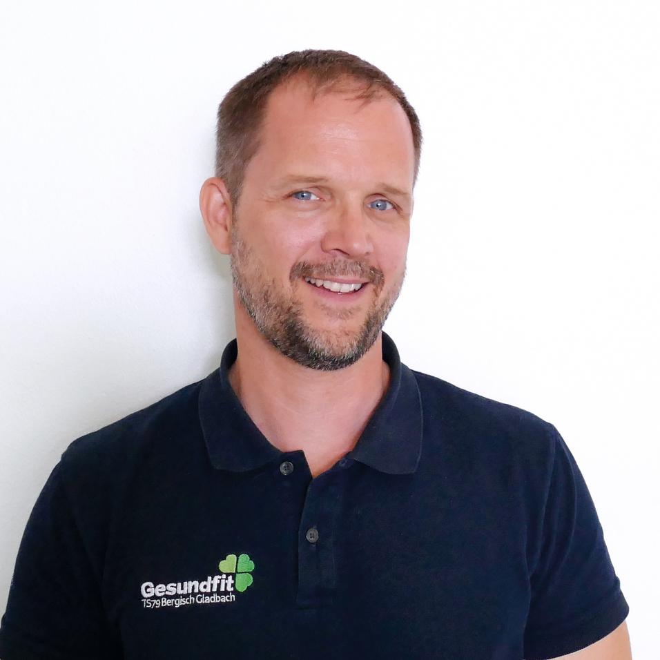 Lächelnder Mann mit kurzer Frisur trägt ein schwarzes Polohemd mit Logo vor weißem Hintergrund.