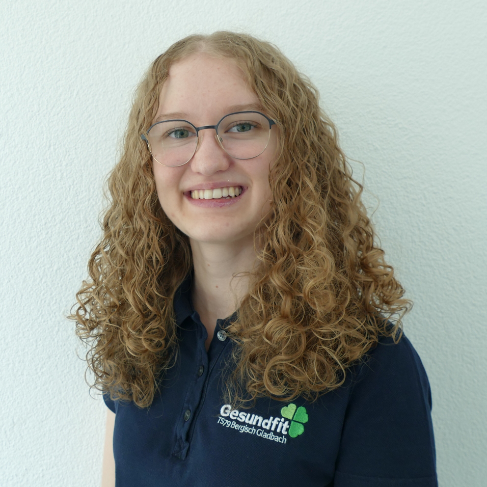 L&auml;chelnde Frau mit lockigem, blondem Haar steht in einem blauen Polohemd vor einer wei&szlig;en Wand.