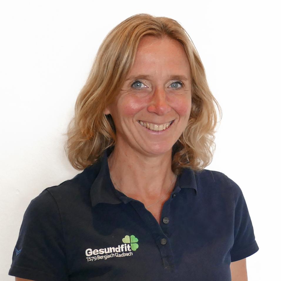 Lächelnde Frau mit mittellangen blonden Haaren, trägt ein schwarzes Polo-Shirt mit dem Aufdruck „Gesundfit“.