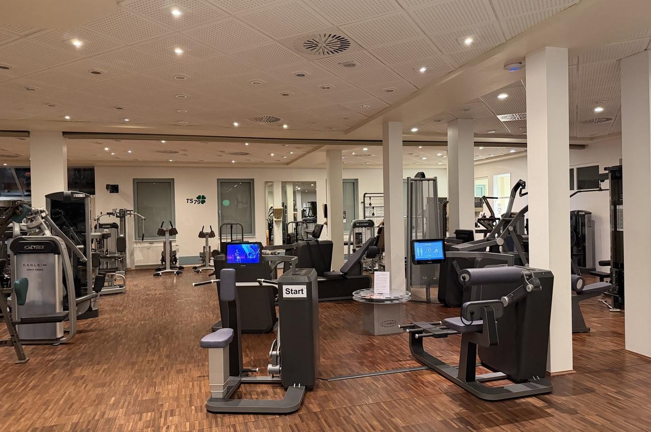 Innenansicht eines Fitnessstudios mit verschiedenen Trainingsgeräten auf einem Holzfußboden und vielen Spiegeln an den Wänden.
