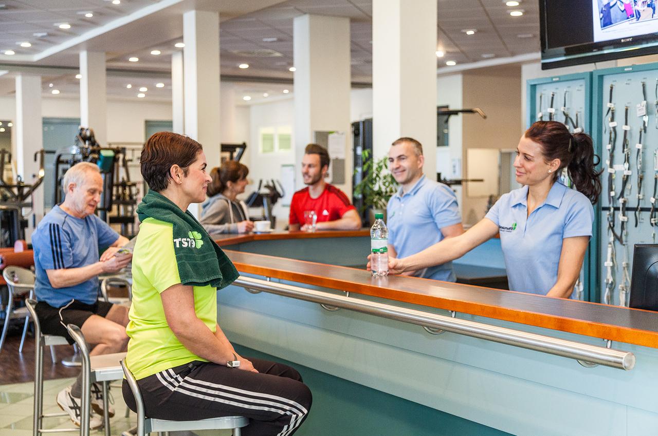 Fünf Personen an einer Fitnessstudio-Bar, eine Frau erhält eine Wasserflasche von einer Mitarbeiterin.