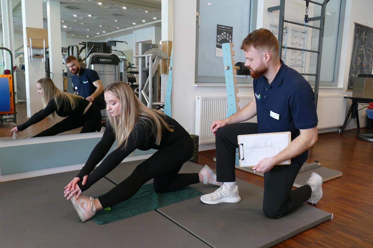 Frau dehnt sich auf einer Trainingsmatte, während ein Trainer mit Notizen daneben kniet und sie anleitet.